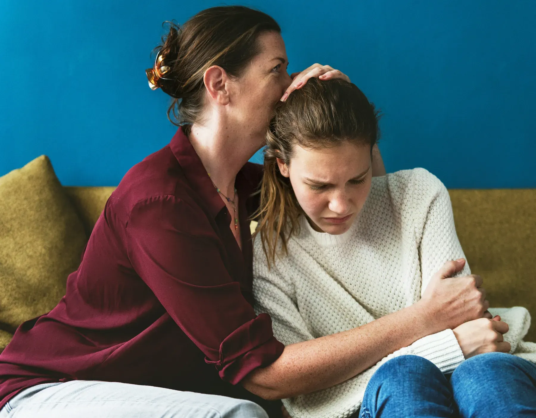 Woman comforting a teenage girl sitting next to her on a couch, with a hand resting on her head