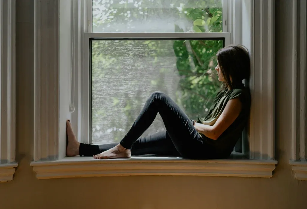 Femme assise sur le rebord intérieur d'une fenêtre, genoux repliés, regardant dehors vers le feuillage