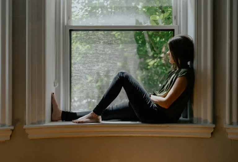 Femme assise sur le rebord intérieur d'une fenêtre, genoux repliés, regardant dehors vers le feuillage