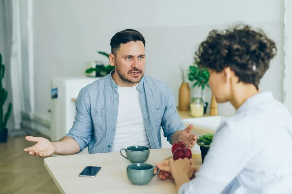 Man speaking with hands spread facing a person seated across a kitchen table