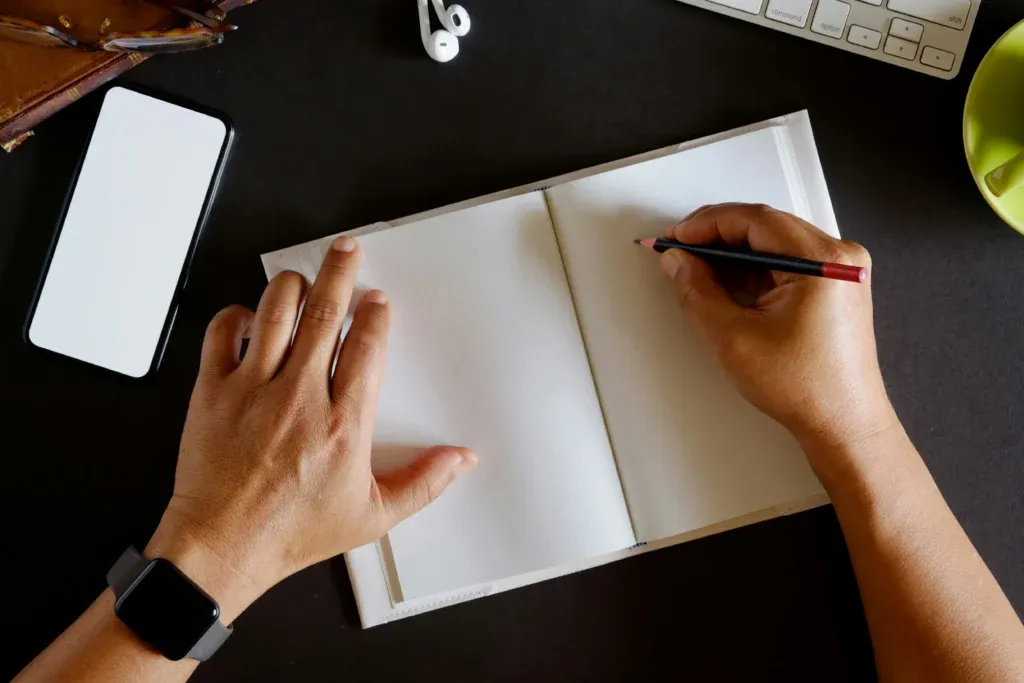 Overhead view of two hands holding a pencil above an open notebook, next to a phone and a keyboard