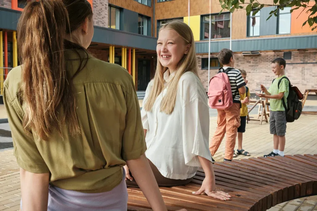 Deux adolescentes assises sur un banc dans une cour d'école, l'une souriant à l'autre, élèves en arrière-plan