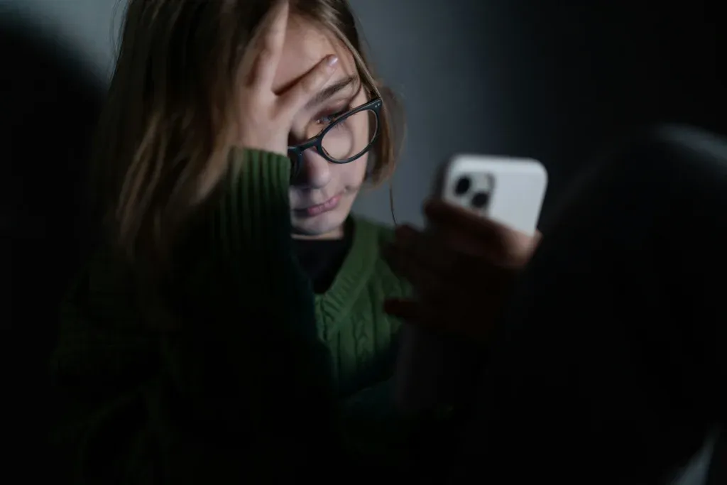 Jeune fille à lunettes dans une pièce sombre, le visage éclairé par l'écran de son téléphone, signes de cyberintimidation